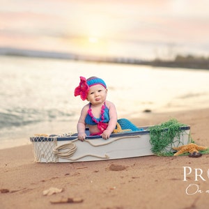 May include: A baby wearing a blue and pink crocheted mermaid costume sits in a small white boat on a sandy beach. The boat is decorated with a net and a starfish. The background is a blurry sunset over the ocean.
