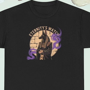 May include: Black t-shirt with a graphic of Anubis, the Egyptian god, and the text "Eternity's Watch". The design features a tan background and purple smoke. The shirt has a crew neck.