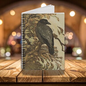 May include: Spiral-bound notebook featuring a detailed illustration of a crow perched on a branch. The artwork is in shades of black, brown, and beige, with a vintage aesthetic. The notebook is open, revealing lined pages.