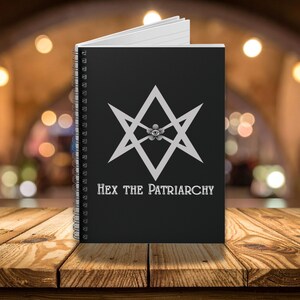 May include: Black spiral-bound notebook with the text "HEX THE PATRIARCHY" in white. The cover features a white geometric design with an eye in the center. The notebook is open, revealing white lined pages.