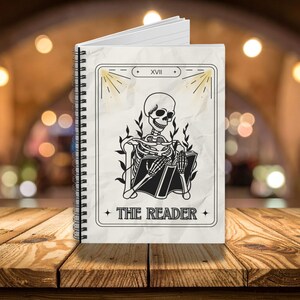 Puede incluir: Cuaderno de espiral con diseño de carta del tarot. La portada muestra una ilustración en blanco y negro de un esqueleto leyendo un libro, con las palabras "THE READER" debajo. El fondo es una imagen borrosa de una mesa de madera.
