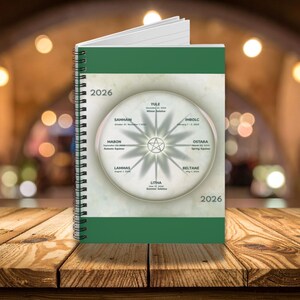 May include: A spiral-bound 2026 planner with a green cover and spine. The cover features a circular design with a star and text indicating seasonal events like Samhain and Litha. The planner is open, revealing lined pages.