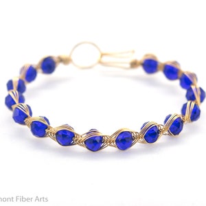Könnte beinhalten: Ein goldfarbenes Drahtarmband mit leuchtend blauen facettierten Perlen. Die Perlen sind gleichmäßig angeordnet und durch ein gewebtes Drahtdesign gehalten. Das Armband hat einen einfachen Verschluss. Das Design ist elegant und alltagstauglich.