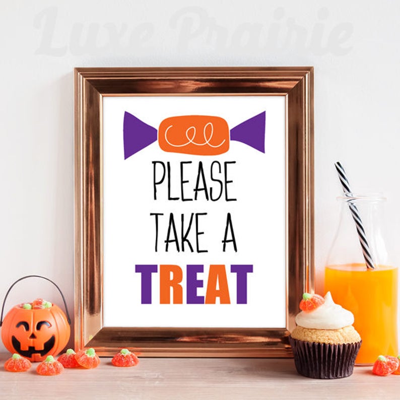 May include: A framed sign with a white background featuring an orange and purple candy graphic at the top and the black and purple text "PLEASE TAKE A TREAT" in the center. The frame is metallic copper and has a rectangular shape.