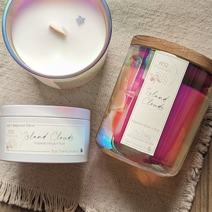 May include: Three candles with iridescent glass and metal containers. The largest candle has a wooden lid and a label that reads "Island Clouds, tropical citrus + fruit, 12 oz". The smallest candle is a tin with a label that reads "Island Clouds, tropical citrus + fruit, 5 oz".