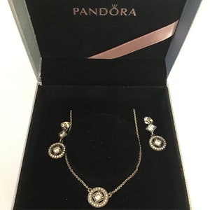 Puede incluir: Una caja blanca con interior negro y un conjunto de collar y pendientes de plata con piedras transparentes. La caja tiene la palabra "PANDORA" impresa en la parte superior.