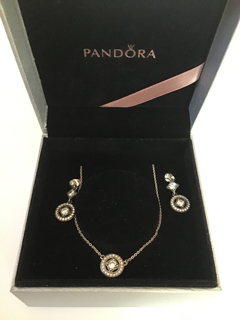 Puede incluir: Una caja blanca con interior negro y un conjunto de collar y pendientes de plata con piedras transparentes. La caja tiene la palabra "PANDORA" impresa en la parte superior.
