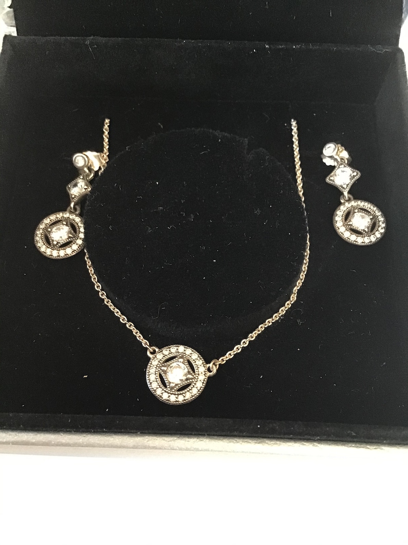 Puede incluir: Un conjunto de collar y pendientes de plata con un colgante redondo y pendientes a juego. El colgante y los pendientes presentan un cristal transparente rodeado de cristales m&aacute;s peque&ntilde;os.