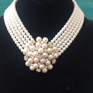 May include: A multi-strand pearl necklace with a central cluster of large pearls and small, sparkling rhinestones. The necklace is displayed on a black surface, showcasing its elegant design and classic appeal.