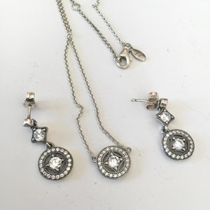 Puede incluir: Un conjunto de collar y pendientes de plata con un colgante circular y un encanto cuadrado colgante, todos adornados con piedras de circonita c&uacute;bica transparentes.