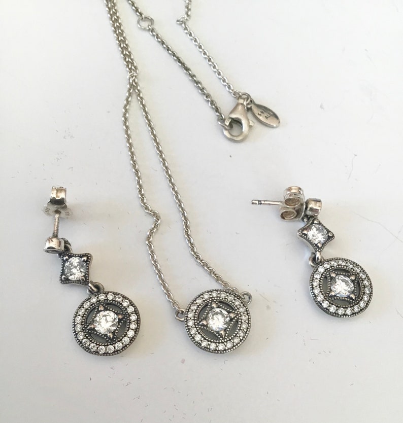 Puede incluir: Un conjunto de collar y pendientes de plata con un colgante circular y un encanto cuadrado colgante, todos adornados con piedras de circonita c&uacute;bica transparentes.