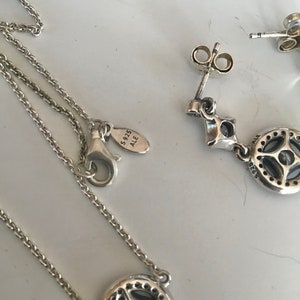 Puede incluir: Un conjunto de collar y pendientes de plata con un colgante redondo que presenta una piedra transparente rodeada de piedras m&aacute;s peque&ntilde;as. El colgante y los pendientes tienen un dise&ntilde;o detallado y el collar tiene una cadena delicada. El cierre del collar tiene una etiqueta que dice "S925 ALE".