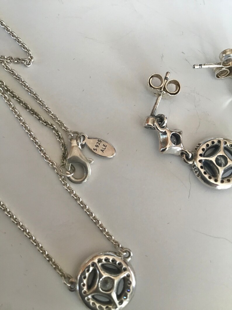 Puede incluir: Un conjunto de collar y pendientes de plata con un colgante redondo que presenta una piedra transparente rodeada de piedras m&aacute;s peque&ntilde;as. El colgante y los pendientes tienen un dise&ntilde;o detallado y el collar tiene una cadena delicada. El cierre del collar tiene una etiqueta que dice "S925 ALE".