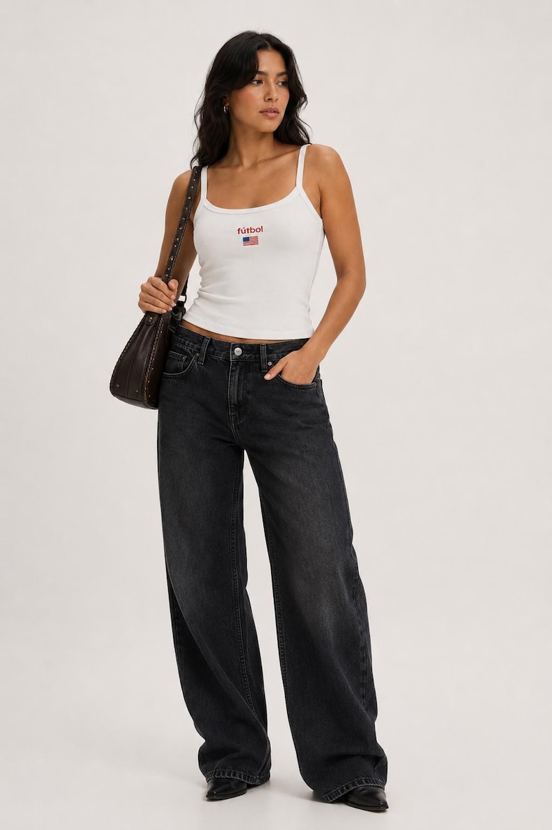 May include: White ribbed tank top with the word "f&uacute;tbol" and a small American flag graphic. The top is paired with dark wash, wide-leg jeans. A brown shoulder bag completes the look.