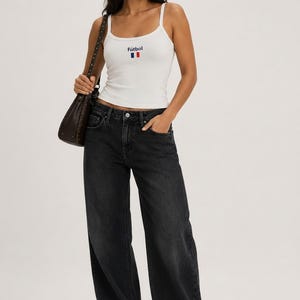 May include: A woman wearing a white tank top with the word "futbol" and a French flag design, paired with black wide-leg jeans. She carries a brown shoulder bag.