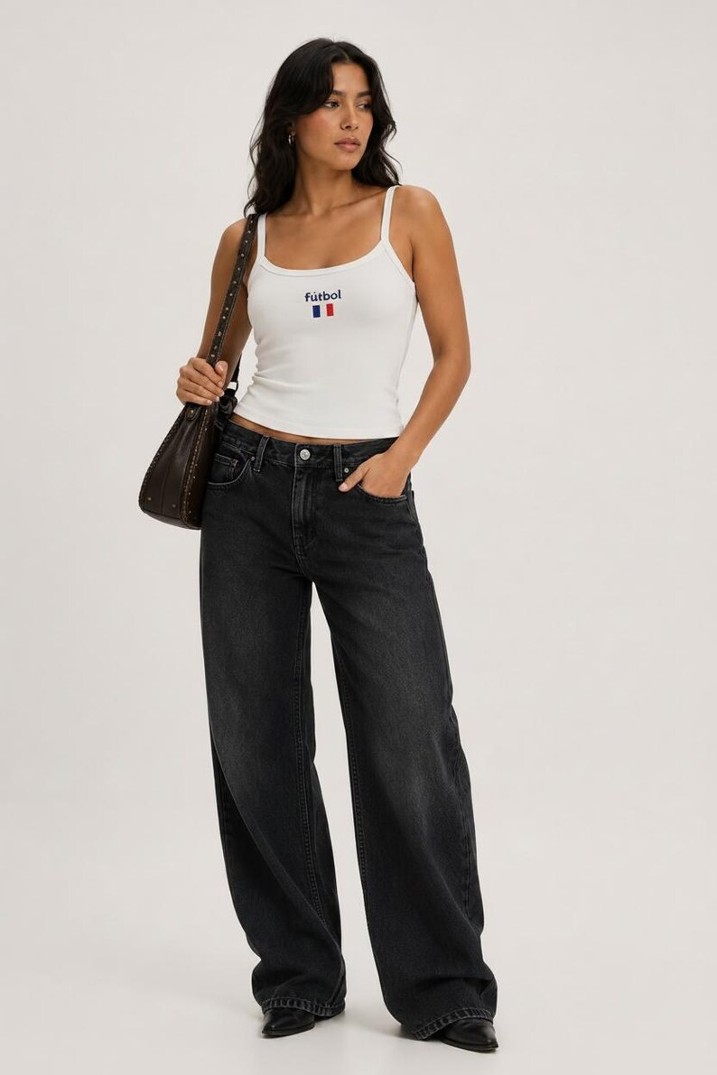 May include: A woman wearing a white tank top with the word "futbol" and a French flag design, paired with black wide-leg jeans. She carries a brown shoulder bag.