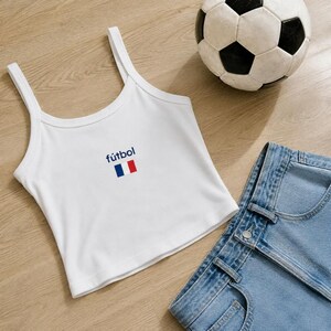 May include: White tank top with the word "f&uacute;tbol" and a French flag design. The top is paired with denim shorts and a soccer ball. The outfit is on a wooden surface.