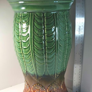 May include: A tall, green ceramic pedestal with a glossy finish. The pedestal features a leaf-like pattern on the body and floral designs at the base. The pedestal is approximately 30 inches tall.