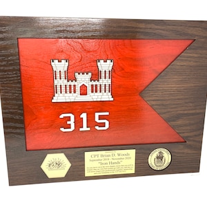 Puede incluir: Placa de madera enmarcada con un dise&ntilde;o de bandera roja, un emblema de castillo blanco y el n&uacute;mero 315. La placa incluye texto y sellos, enmarcada en madera marr&oacute;n oscuro. El texto dice "CPT Brian D. Woods" y "Iron Hands".