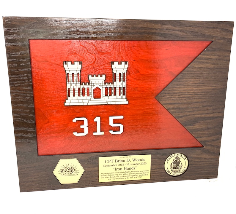 Puede incluir: Placa de madera enmarcada con un dise&ntilde;o de bandera roja, un emblema de castillo blanco y el n&uacute;mero 315. La placa incluye texto y sellos, enmarcada en madera marr&oacute;n oscuro. El texto dice "CPT Brian D. Woods" y "Iron Hands".
