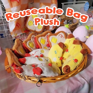 Puede incluir: Una cesta de mimbre llena de juguetes de peluche de comida, incluyendo tostadas, plátanos y sushi. Los juguetes son de colores brillantes con costuras detalladas. El texto "Reusable Bag Plush" está en letras naranjas.