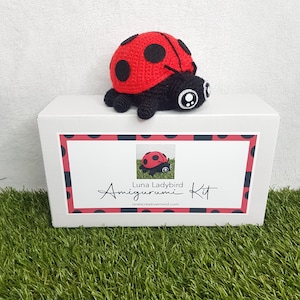 May include: A white box with a red and black polka dot pattern. The box has a label that says "Luna Ladybug Amigurumi Kit" and "onecreativemind.com". There is a small red and black ladybug amigurumi on top of the box.