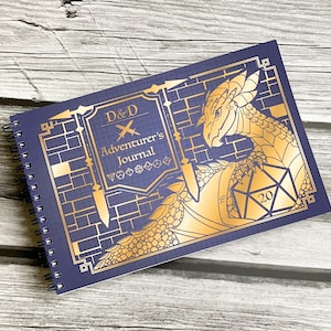 May include: A blue spiral bound journal with a gold dragon design and the text "D&D Adventurer's Journal". The dragon is holding a gold 20-sided die.