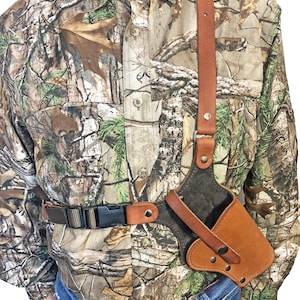 May include: A brown leather shoulder holster with a black fabric lining. The holster is worn over a camouflage shirt and secured with a brown leather belt.