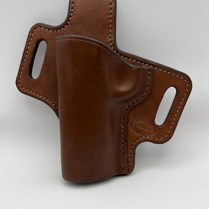 May include: A brown leather holster featuring a molded shape for a firearm, reinforced stitching along the edges, and two belt slots. An oval stamp on the lower right side displays the text Western Images Colorado Saddlery, Inc.