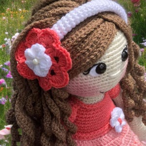 Pode incluir: Boneca de croch&ecirc; feita &agrave; m&atilde;o com cabelo castanho encaracolado, vestindo um vestido rosa e uma faixa branca. Uma flor vermelha e branca adorna o cabelo da boneca. A boneca tem olhos grandes e pretos e uma express&atilde;o doce.