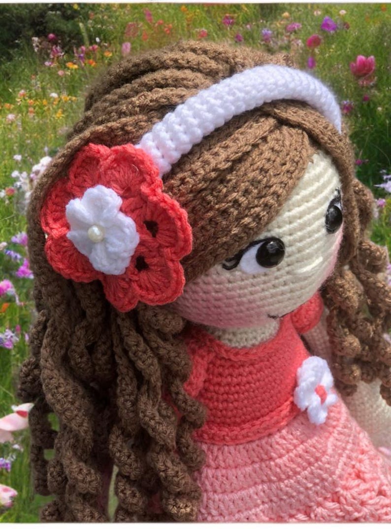 Pode incluir: Boneca de croch&ecirc; feita &agrave; m&atilde;o com cabelo castanho encaracolado, vestindo um vestido rosa e uma faixa branca. Uma flor vermelha e branca adorna o cabelo da boneca. A boneca tem olhos grandes e pretos e uma express&atilde;o doce.