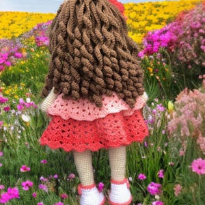 Pode incluir: Boneca de croch&ecirc; feita &agrave; m&atilde;o com cabelo encaracolado castanho, uma flor vermelha e um vestido rosa e vermelho em camadas. A boneca est&aacute; usando sapatos brancos com detalhes vermelhos e est&aacute; em um campo de flores.