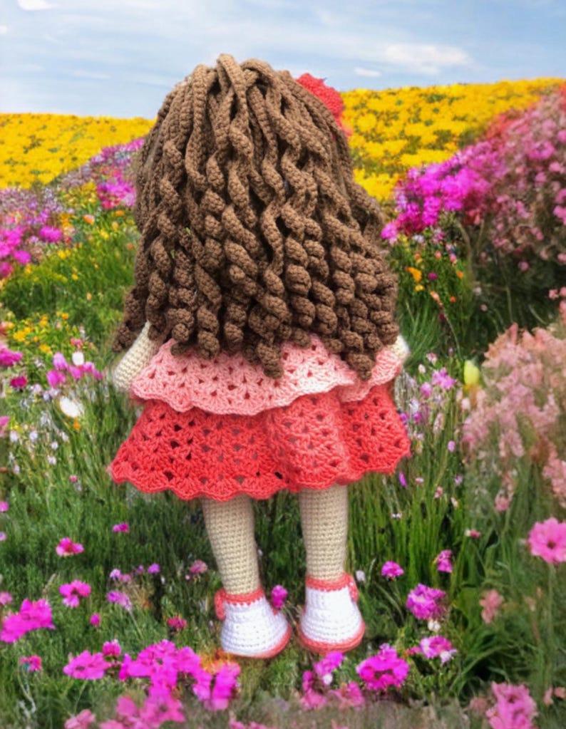 Pode incluir: Boneca de croch&ecirc; feita &agrave; m&atilde;o com cabelo encaracolado castanho, uma flor vermelha e um vestido rosa e vermelho em camadas. A boneca est&aacute; usando sapatos brancos com detalhes vermelhos e est&aacute; em um campo de flores.