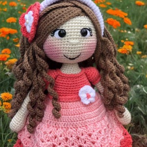 Pode incluir: Boneca de croch&ecirc; feita &agrave; m&atilde;o com cabelo castanho encaracolado, vestindo um vestido rosa com uma flor branca. A boneca tem uma faixa branca e uma flor na lateral da cabe&ccedil;a. Flores laranja ao fundo.
