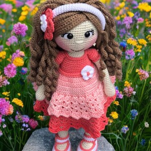 Pode incluir: Boneca de croch&ecirc; feita &agrave; m&atilde;o com cabelo castanho encaracolado, vestindo um vestido rosa com um detalhe de flor branca. A boneca tem uma faixa branca e est&aacute; em p&eacute; sobre uma rocha em um campo de flores coloridas. Os sapatos da boneca s&atilde;o brancos com detalhes em rosa.