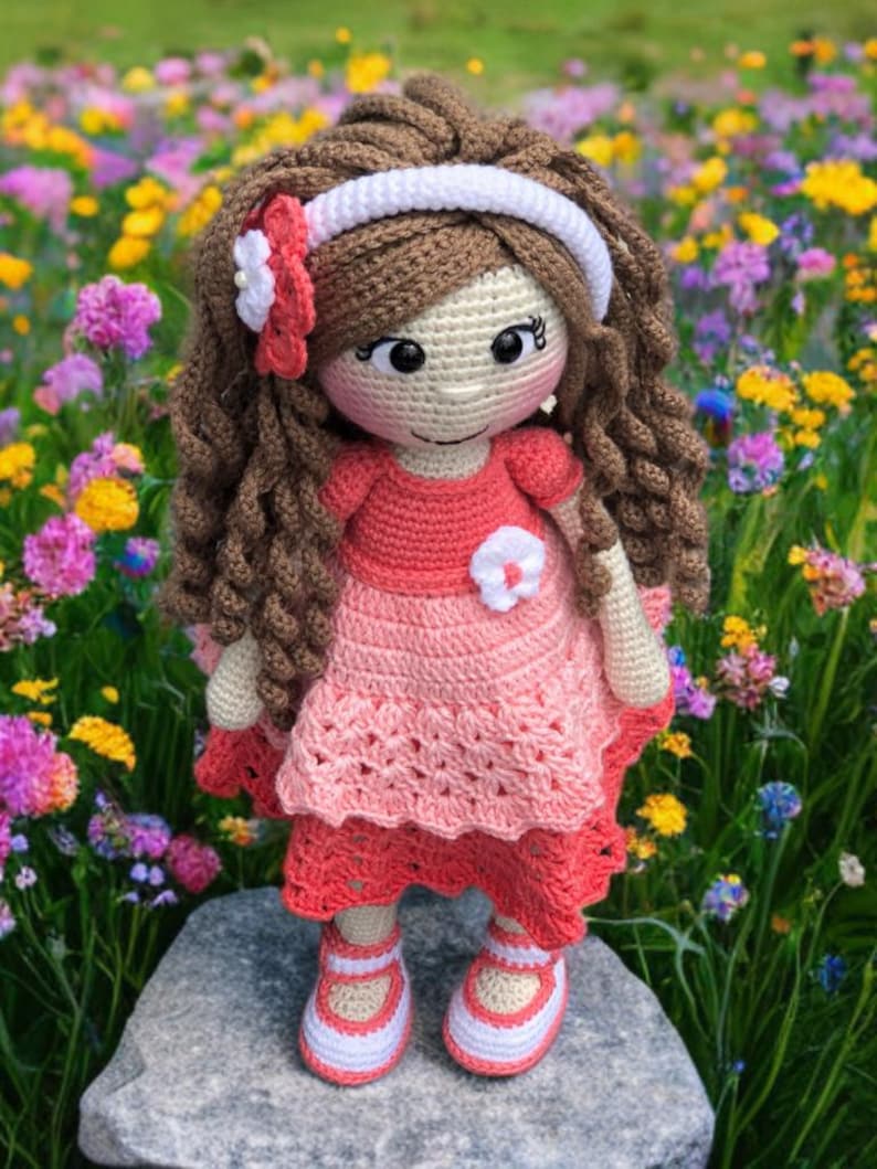 Pode incluir: Boneca de croch&ecirc; feita &agrave; m&atilde;o com cabelo castanho encaracolado, vestindo um vestido rosa com um detalhe de flor branca. A boneca tem uma faixa branca e est&aacute; em p&eacute; sobre uma rocha em um campo de flores coloridas. Os sapatos da boneca s&atilde;o brancos com detalhes em rosa.