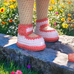 Pode incluir: Botinhas de beb&eacute; de croch&eacute; em branco e coral. As botinhas t&ecirc;m um estilo Mary Jane com uma tira e um pequeno detalhe floral. As pernas s&atilde;o de croch&eacute; em bege. As botinhas est&atilde;o sobre uma superf&iacute;cie de pedra com um fundo desfocado de flores.