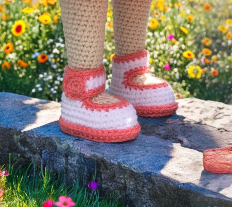 Pode incluir: Botinhas de beb&eacute; de croch&eacute; em branco e coral. As botinhas t&ecirc;m um estilo Mary Jane com uma tira e um pequeno detalhe floral. As pernas s&atilde;o de croch&eacute; em bege. As botinhas est&atilde;o sobre uma superf&iacute;cie de pedra com um fundo desfocado de flores.