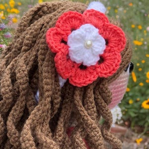 Pode incluir: Boneca de croch&ecirc; feita &agrave; m&atilde;o com cabelo de fio marrom e um acess&oacute;rio de cabelo de flor coral e branco. O cabelo da boneca &eacute; estilizado em mechas longas e encaracoladas. A flor tem um centro de p&eacute;rola e &eacute; presa a uma faixa branca.