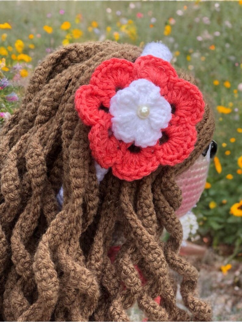 Pode incluir: Boneca de croch&ecirc; feita &agrave; m&atilde;o com cabelo de fio marrom e um acess&oacute;rio de cabelo de flor coral e branco. O cabelo da boneca &eacute; estilizado em mechas longas e encaracoladas. A flor tem um centro de p&eacute;rola e &eacute; presa a uma faixa branca.