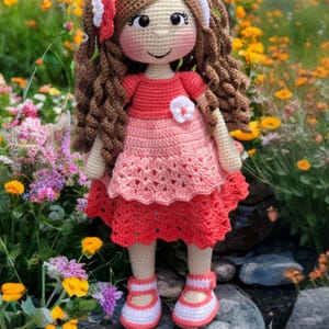 Pode incluir: Boneca de croch&ecirc; feita &agrave; m&atilde;o com cabelo castanho encaracolado, vestida com um vestido vermelho e rosa com uma flor branca. A boneca tem uma faixa branca e sapatos a condizer. Est&aacute; em p&eacute; sobre uma rocha num jardim florido.