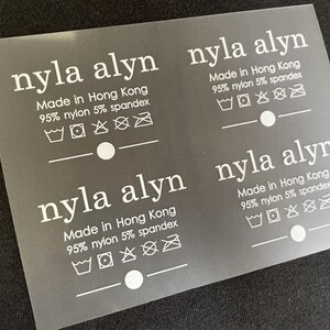 K&ouml;nnte beinhalten: Drei wei&szlig;e Pflegeetiketten mit dem Text "nyla alyn" und "Made in Hong Kong 95% nylon 5% spandex". Jedes Etikett enth&auml;lt eine Reihe von Waschanweisungen.