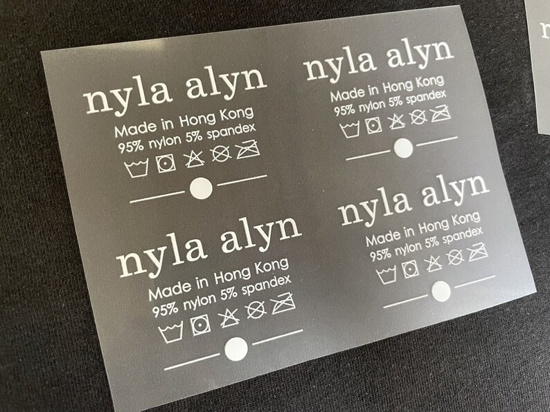 K&ouml;nnte beinhalten: Drei wei&szlig;e Pflegeetiketten mit dem Text "nyla alyn" und "Made in Hong Kong 95% nylon 5% spandex". Jedes Etikett enth&auml;lt eine Reihe von Waschanweisungen.