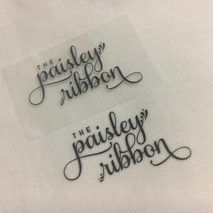 K&ouml;nnte beinhalten: Zwei transparente Etiketten mit schwarzem Text, der in einer kursiven Schrift "The paisley ribbon" lautet.