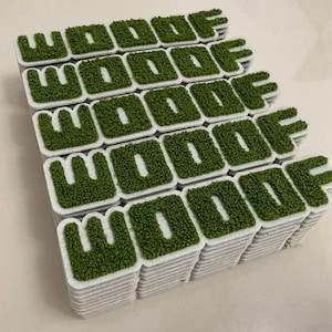 May include: A stack of rectangular white patches, each featuring the word WOOOF in raised, textured green material. The letters have a fuzzy, moss-like appearance and are centred on each individual white rectangular base in this collection.