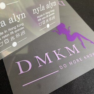 K&ouml;nnte beinhalten: Ein wei&szlig;es Etikett mit dem Text "nyla alyn" und "Made in Hong Kong 95% nylon 5% spandex" mit Pflegehinweisen. Das Etikett befindet sich auf einem lila und wei&szlig;en Logo mit der Aufschrift "DMKM" und "DO MORE KNOW MORE".