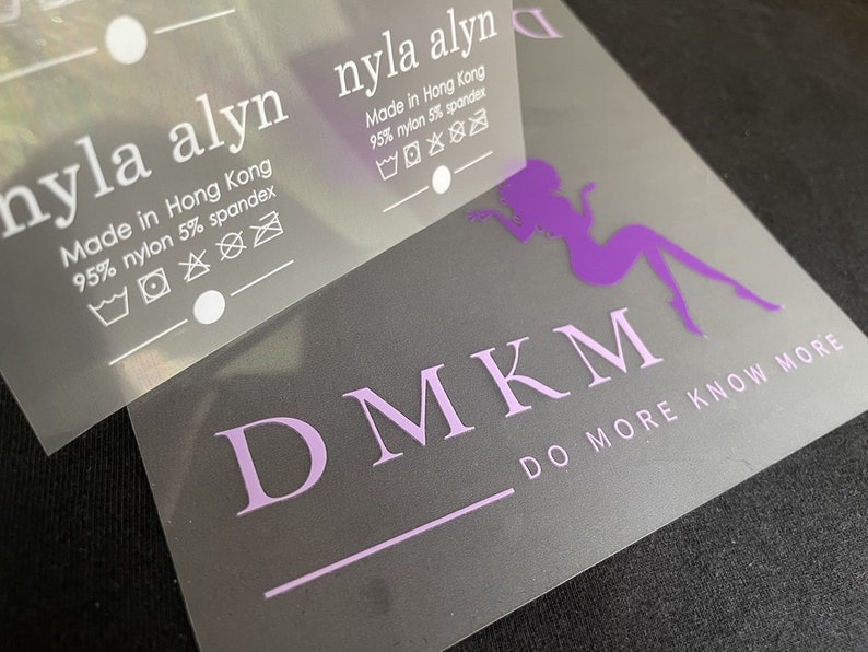 K&ouml;nnte beinhalten: Ein wei&szlig;es Etikett mit dem Text "nyla alyn" und "Made in Hong Kong 95% nylon 5% spandex" mit Pflegehinweisen. Das Etikett befindet sich auf einem lila und wei&szlig;en Logo mit der Aufschrift "DMKM" und "DO MORE KNOW MORE".