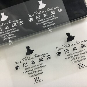 K&ouml;nnte beinhalten: Ein transparentes Kleidungsetikett mit schwarzem Text und Symbolen. Das Etikett lautet "Sea Waters Designs" und enth&auml;lt Pflegehinweise, Gr&ouml;&szlig;e und Stoffzusammensetzung. Die Stoffzusammensetzung ist 80 % Nylon und 20 % Elasthan oder 90 % Polyester und 10 % Elasthan. Die Gr&ouml;&szlig;en sind S und XL.