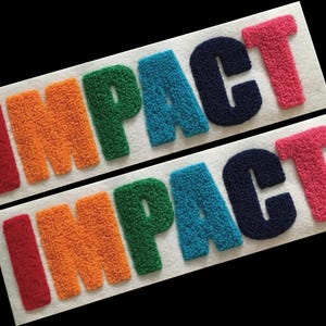 Puede incluir: Dos tiras de tela blanca que presentan la palabra IMPACT bordada en un estilo de aguja perforada texturizado y multicolor. Las letras son de color rojo, naranja, verde, azul, azul oscuro y rosa, dispuestas en una secuencia horizontal.