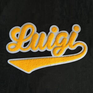 Puede incluir: Un parche de chenilla amarillo con el nombre Luigi en una fuente cursiva. Las letras tienen una superficie texturizada y difusa, delineadas con un borde blanco grueso, con una larga cola amarilla que se extiende bajo el texto.